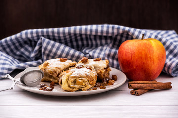 Slices of homemade Apple pie strudel in a plate with ingredients on a rustic white wooden table. Pie with apples, raisins and cinnamon, sweet pastries