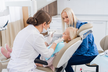 Fototapeta premium A child with a mother at a dentist's appointment. The girl lies in the chair with her mouth open, with her mother behind. The doctor is doing her job