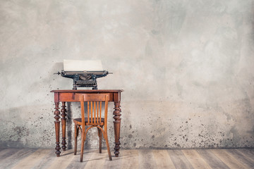 Old typewriter with sheet of paper on retro oak desk and aged chair on wooden floor front grunge concrete wall background. Writer's classic workplace concept in loft room. Vintage style filtered photo