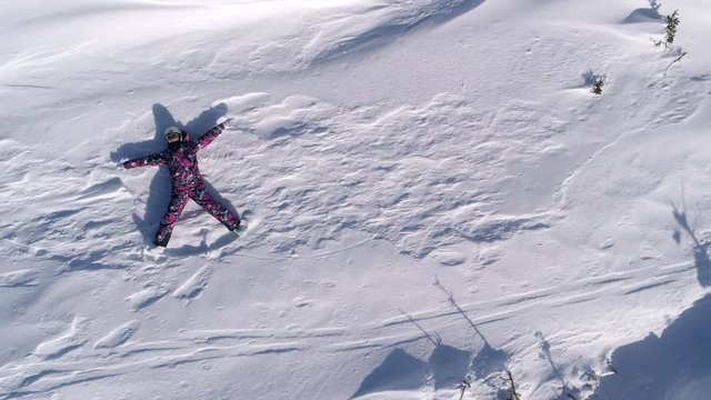 Top aerial view Footage: a girl in a pink jumpsuit depicts the figure of a snow angel. a skier lies on a snow covered field in the mountains and waves her arms and legs surrounded by snow clouds