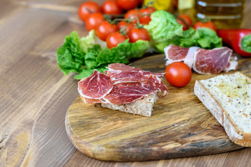 White bread and slices of smoked pork ham on the wooden kitchen board. Vegetables, green and olive oil on the kitchen table. Healthy tasty food with minerals and vitamins.