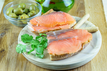 Sandwiches with red fish salmon on the wooden plate, olives and olive oil closeup. Tasty mediterranean snack food.