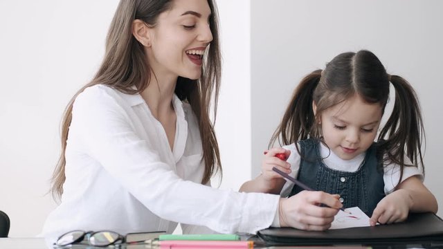 A mother is painting with her daughter. The woman is helping the kid to color the picture. The kid is happy to do the activity with her mother.