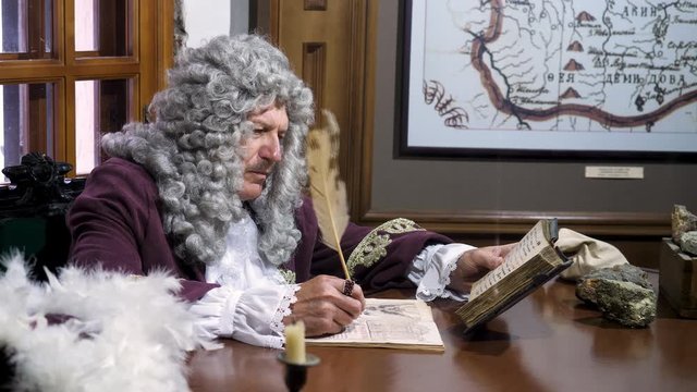 Retro baroque man with white wig holding a quill pen, sitting at antique table and thinking about paper documents, history concept. Stock footage. Reconstruction of historical events at the museum.
