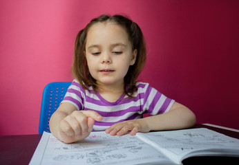 A cute little girl is sitting at a table and doing her homework. Pink background. Distance learning at home