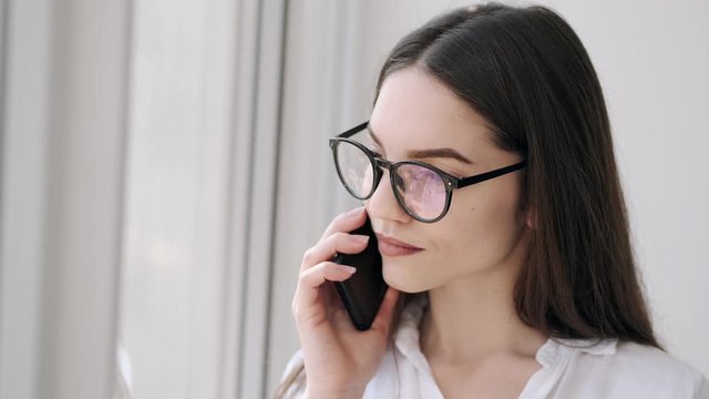 A beautiful girl in glasses is talking on the phone. She is standing near the window an smiling while the communication.