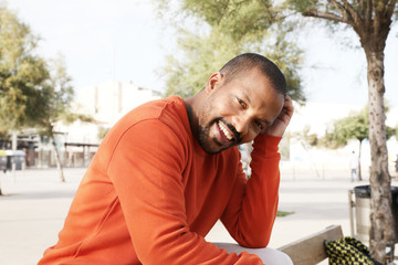 Happy African American man sitting on bench in park wearing casual clothing. Horizontal.