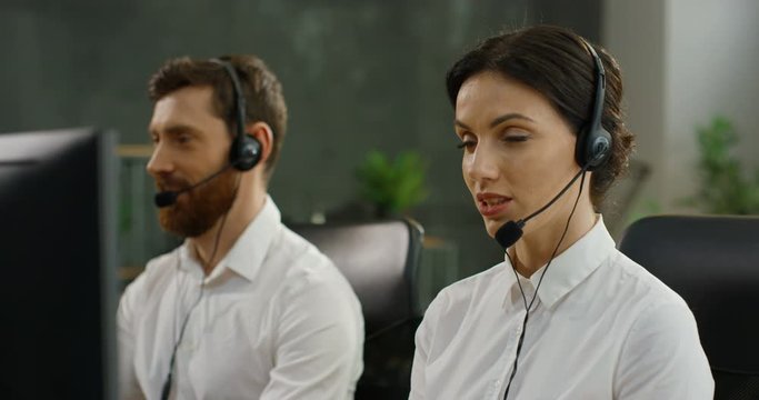 Close up of Caucasian beautiful woman in headset chatting with customer at computer and solving problem. Female colleagues in call center office. Young male co-worker talking with client on background