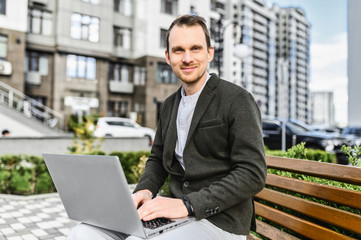 A young happy man in smart casual uses laptop for remote work outdoors. He sits on the bench among cityscape, looking at camera with smile