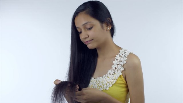A young girl holds her long hair looking at the damage - damaged hair concept. Pretty Indian female is very sad and upset while observing the split ends and frizzy texture of her long black hair