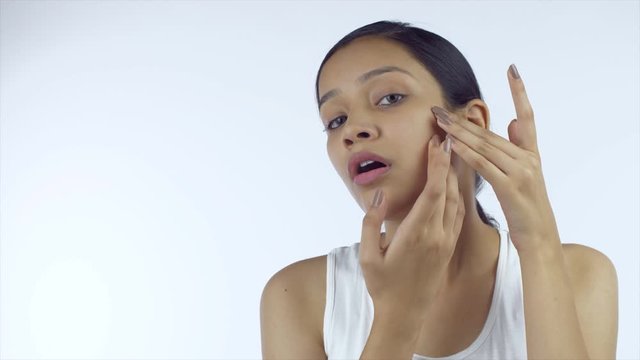 Unhappy young female looking at her skin condition while touching her face - Acne and Pimple. Beautiful Indian girl with no makeup checking her face while looking at the camera against the white ba...
