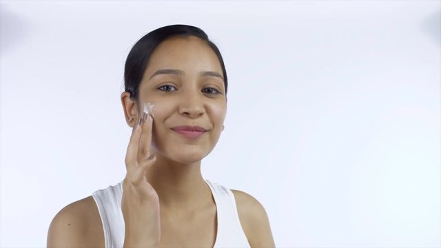 Young girl massaging little amount of cream gently on her face - beauty concept. Closeup shot of a beautiful female with no makeup applying moisturizer on her cheeks against the white background