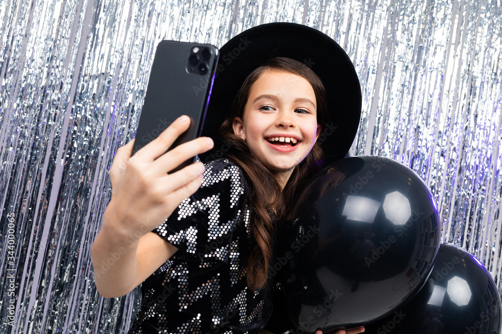 Wall mural young girl on holiday takes a photo on a smartphone among black balloons on a shiny background