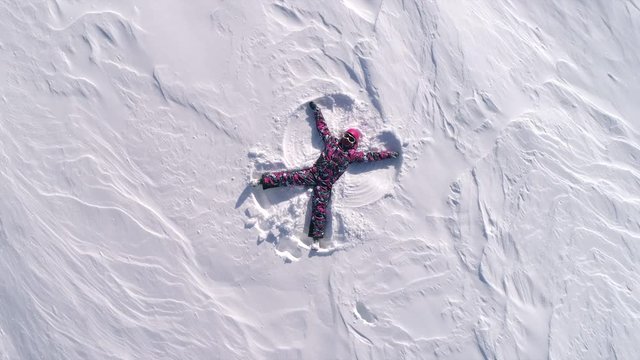 AERIAL UP spin: snow angels in winter. a girl in a pink ski suit and helmet with glasses depicts a snow angel, waving her arms and legs while lying on the fresh white snow. winter outdoor activity. 4k