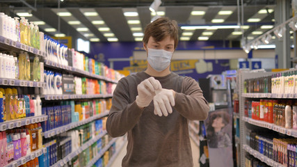 Coronavirus pandemic protection. Man in a medical mask puts on rubber gloves in a supermarket between shelves with goods in slow motion.