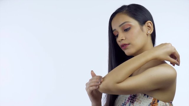 Side view of a young woman applying moisturizer on her elbow - skincare concept. Beautiful Indian girl with open black hair rubbing cream on her elbow to keep her skin moisturized - white background