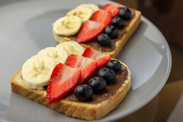 Sandwiches with fruits on a grey plate. Concept of delicious and healthy breakfast. Toasts with chocolate topping, banana, strawberry and blueberry.