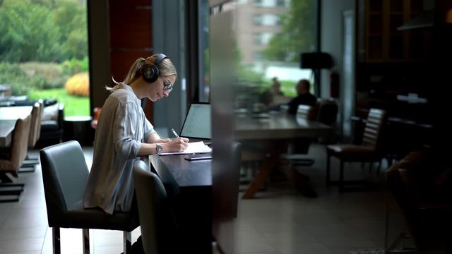 Female employee having overtime work sitting at table and editing financial reports connected to 4g wireless on laptop computer for listening music via electronic headphones for noise cancellation
