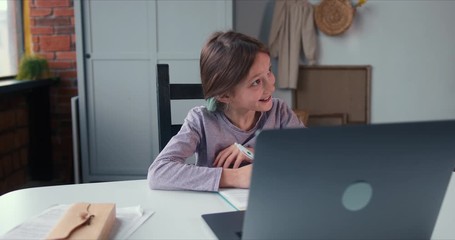 Cute smiling Caucasian teen age girl writes down tasks talking to remote tutor via laptop webcam app on self isolation.
