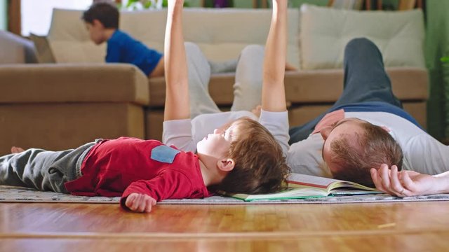 Small cute boy with their parents spending time together while laying down on the floor other big boy sitting on the sofa and playing a game on the laptop