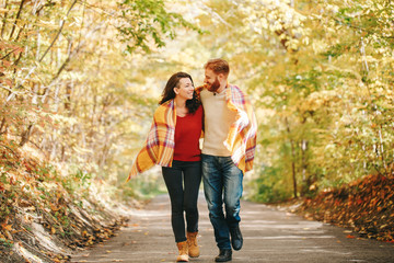 Beautiful couple man woman in love. Smiling laughing boyfriend and girlfriend wrapped in yellow blanket walking in a park on autumn fall day. Togetherness and happiness. Authentic real people.