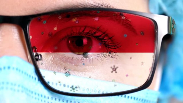 closeup, eye, part of doctor face in medical mask, glasses, which painted in colors of Monaco flag. Many viruses, germs moving on glass.State interests in vaccines, drugs invention, pathogenic viruses