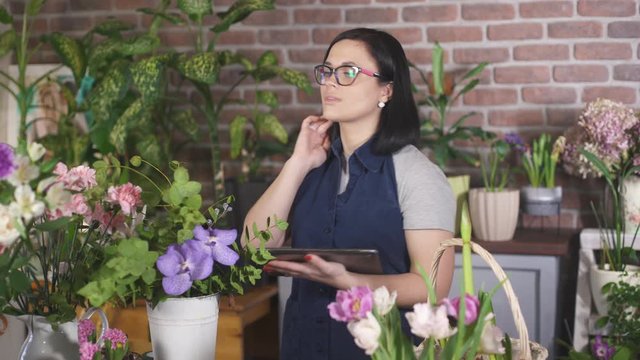 Brunette girl in glasses uses a tablet. Woman florist uses technology to count flowers in store. Female owner a flower shop uses gadgets in their business. Businesswoman small business owner.