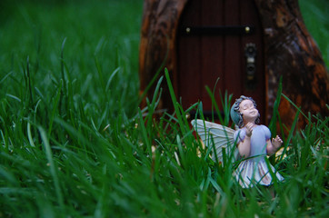 Sweet fairy statue stands in fairy garden surrounded by green grass. Fairy princess is in front of door to magical fantasy world. She is beautiful, mystical, and delicate.