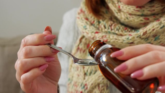 people, health and cold concept - sick woman in blanket and scarf drinking cough syrup at home