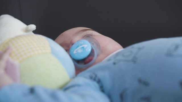 Cute Caucasian Male Newborn Baby Have Fun With A Ball On The Bed In A Room. Then Spit Out A Pacifier. Interior Scene. Close Up Shot.