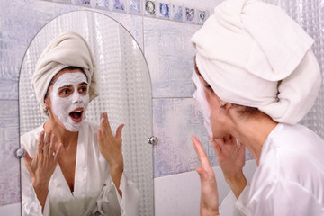 Beautiful brunette woman at home in pajama and towel on her head put white mask on her face at bathroom
