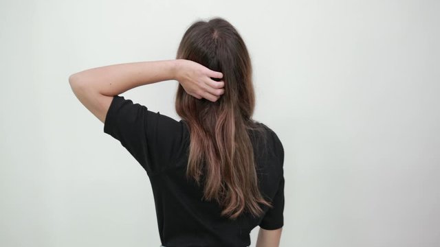 Cute young brunette woman in black t-shirt on gray background, the girl turned her back to camera and holds her hand in long hair