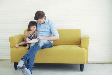 Young handsome Father hugs his little daughter while they are drawing and coloring on a book together on the yellow sofa at home