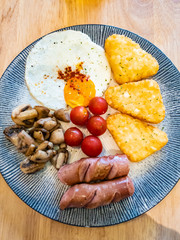 Western style breakfast on a blue circular plate. Consists of sunny side up, beef sausage, cherry tomatoes, hash brown, and grilled champignon mushroom.