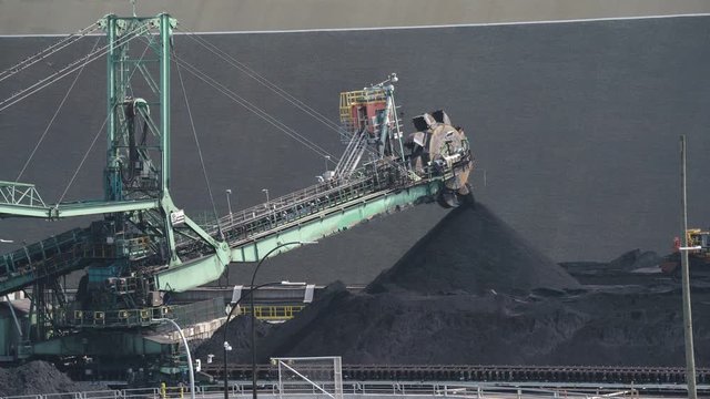 A time lapse of a large stacker machine piling coal in North Vancouver port while tiny-looking bulldozer flattens out piles 