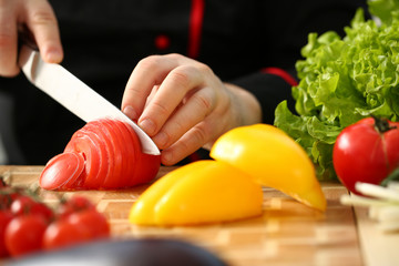 Cook holds knife in hand and cuts on cutting board red tomatoes for salad or fresh vegetable soup with vitamins. Raw food and vegetarian recipe book in modern society popular concept.