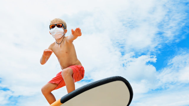 Young funny surfer wearing sunglasses, protective face mask ride on surfboard. Summer beach tours, cruises cancelled due to coronavirus COVID-19 epidemic. Safe travel destinations for family vacation.