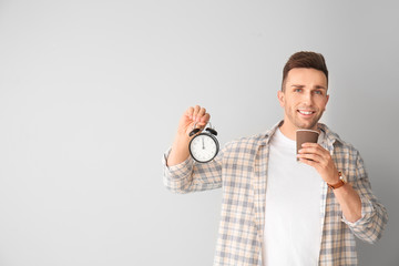 Young man with alarm clock drinking coffee on light background
