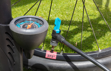 A close up of a pressure gauge in front of a mountain bike tire. The hose of an air pump is connected. The valve is open and air is pumped up.