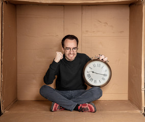 young unemployed student sits in a tight cardboard box, the idea of unemployment without experience, the clock in the hands of a symbol of the passage of time