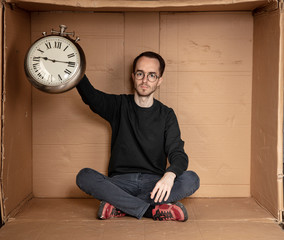 young unemployed student sits in a tight cardboard box, the idea of unemployment without experience, the clock in the hands of a symbol of the passage of time