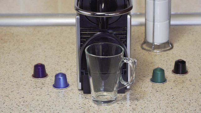 Creating espresso on an automatic machine using aluminum capsules. Female at kitchen sink adds a pod on top of coffeemaker and makes hot dripping coffee with rich cream on a glass cup.