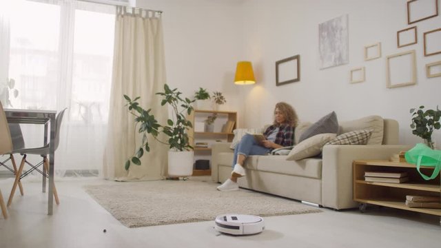 Medium shot of young woman sitting on couch and reading book while white robot vacuum cleaner working in living room