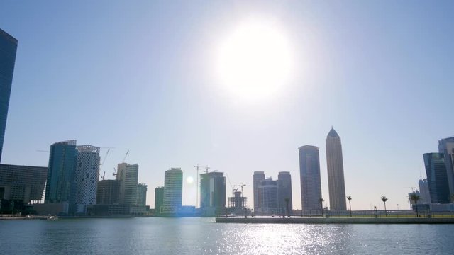 Low angle view of lake water, bright white sun and city skyscrapers on horizon, Dubai