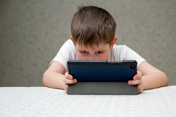 The boy plays on the digital tablet. Boy with tablet sitting on chair, on home interior background. Happy childhood. Positive emotions and energy