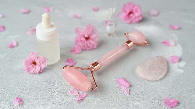 rose quartz roller and facial serum bottle lie on white background with pastel pink flowers and petals close view