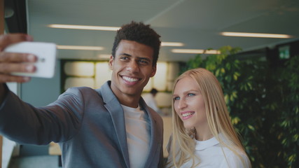 Cheerful black man with laughing blond woman taking selfie with smartphone in modern office having fun.