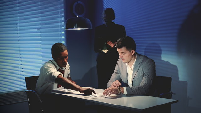 Medium Shot Of African Detective Questioning Suspect Caucasian Man In Interrogation Room. Female Assistant Making Notes Of The Interrogation.