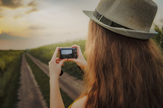 A young woman with red mirrorless camera is taking photos of a beautiful summer landscape. A woman photographer is walking by the field with her camera