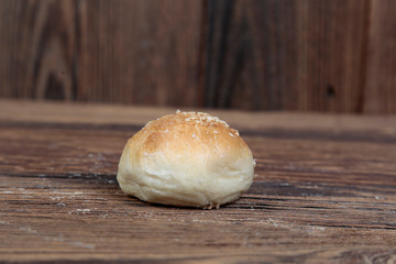 Side view of a handmade, fresh, round bun sprinkled with white sesame seeds on a wooden, brown, rustic table. 